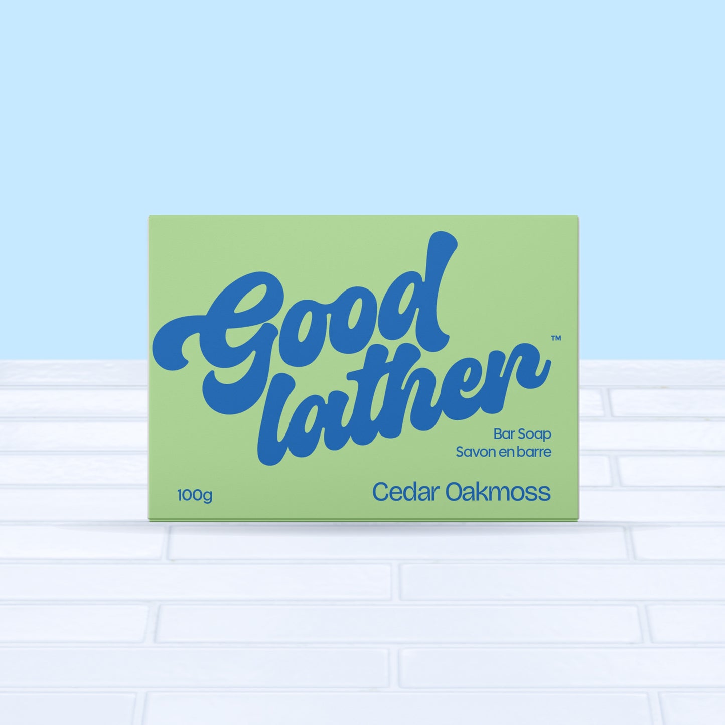 A green and bright blue box of Goodlather Cedar Oakmoss Bar Soap rests on white tile with a light blue background. The Canadian-made packaging features bold blue text and holds 100g of sustainable product.