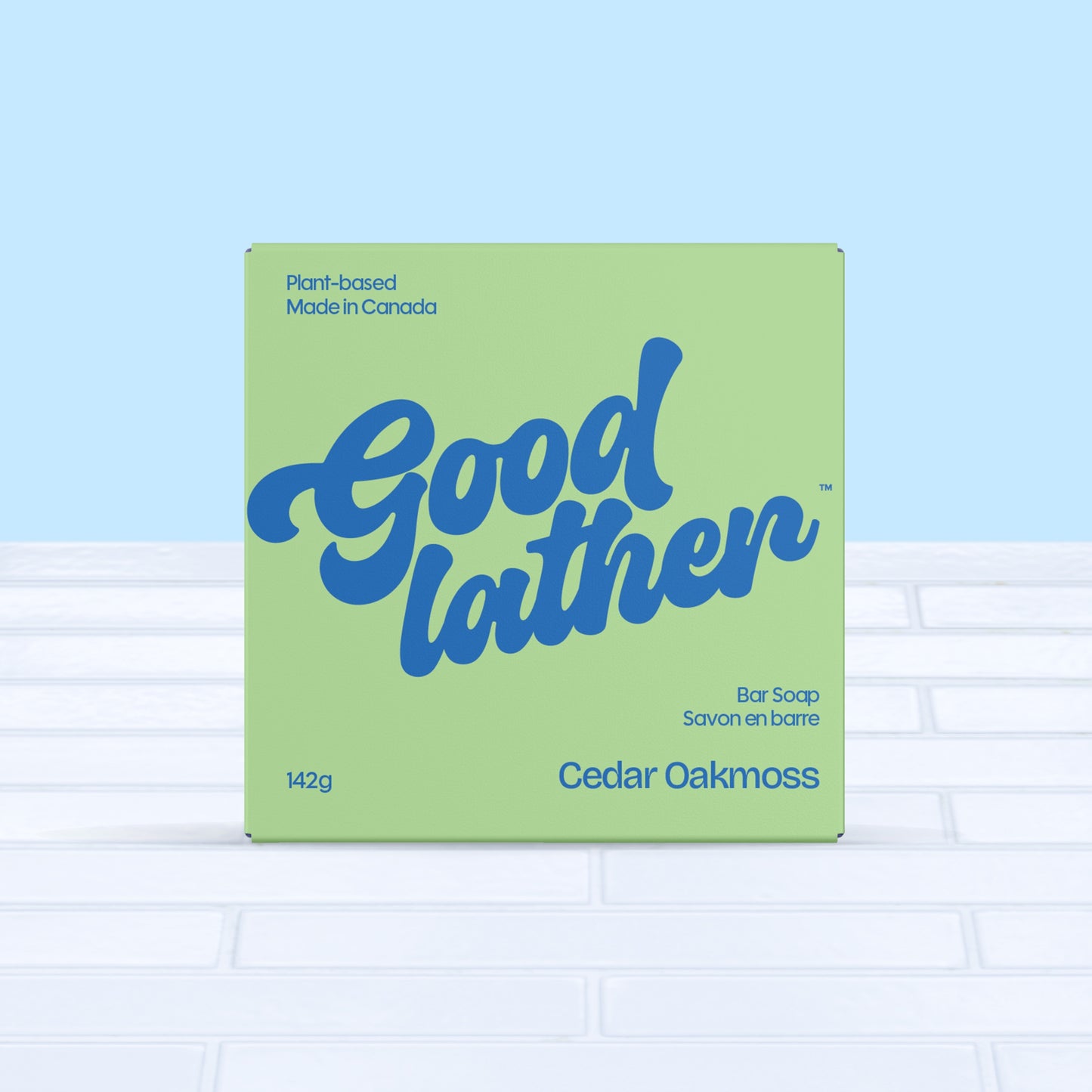 A green and bright blue box of Goodlather Cedar Oakmoss Bar Soap rests on white tiles with a light blue background. This plant-based, Canadian-made bar features a cedar oakmoss scent and supports sustainable practices.