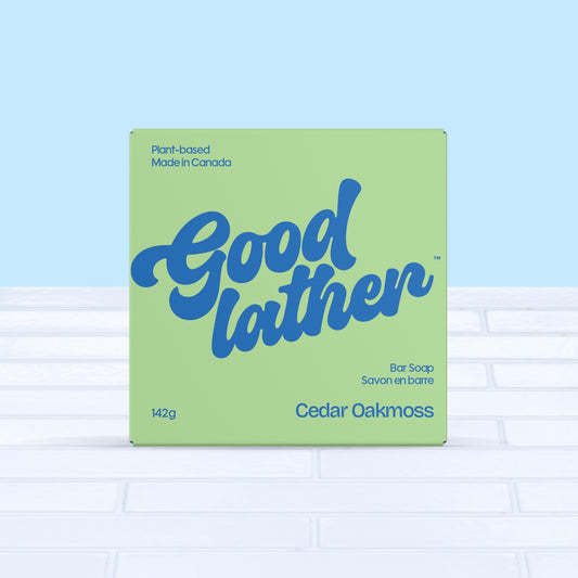 A green and bright blue box of Goodlather Cedar Oakmoss Bar Soap rests on white tiles with a light blue background. This plant-based, Canadian-made bar features a cedar oakmoss scent and supports sustainable practices.