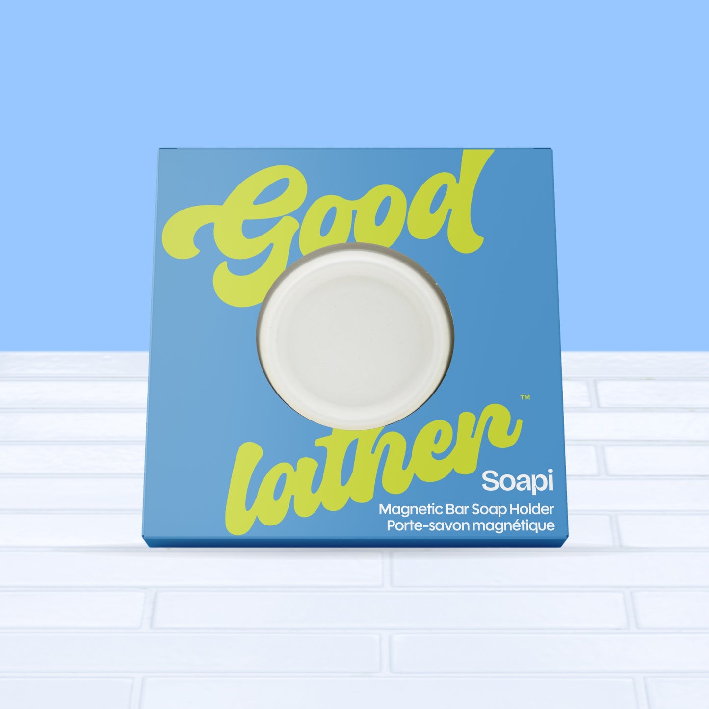 A white Goodlather Soapi Magnet Holder displays the round soap holder on a tiled surface with a blue background—ideal for modern, eco-conscious homes.